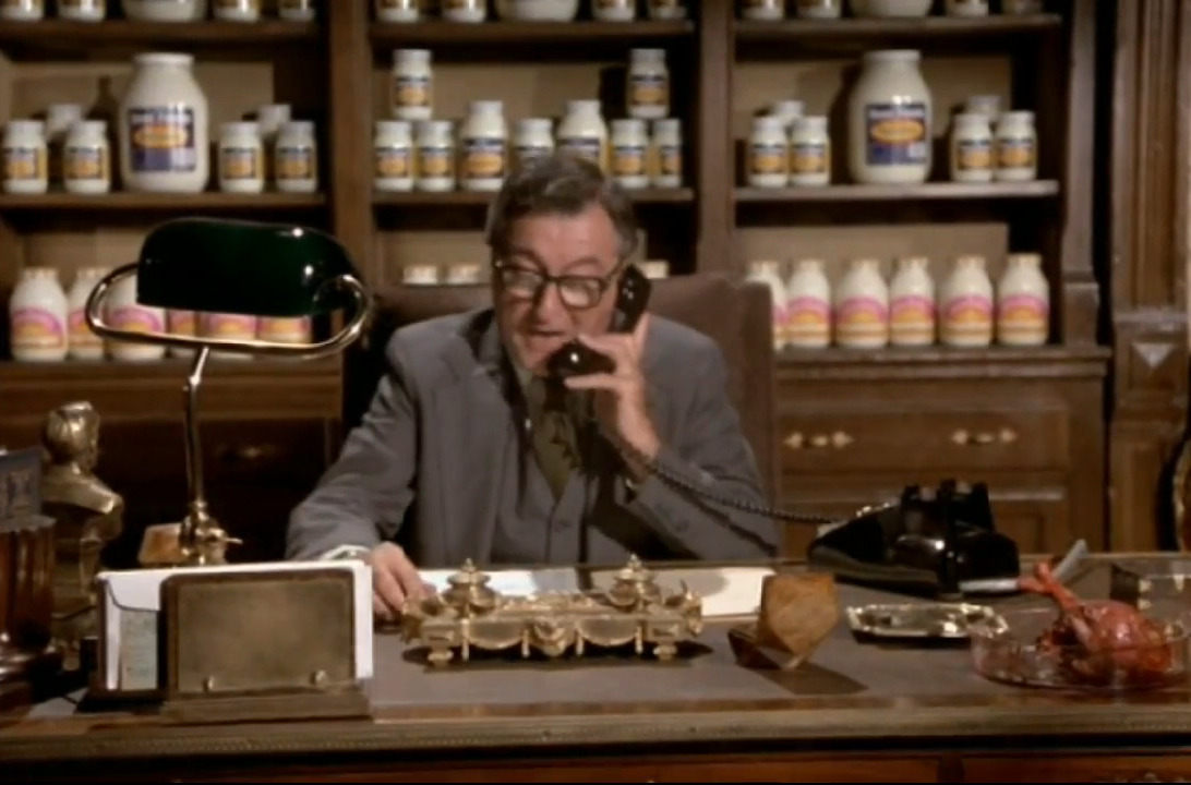 a still from the movie 'Airplane!' where somebody at 'mayo clinic' is sitting at an office desk using a phone. there are bookshelves behind him filled with jars of mayonnaise.