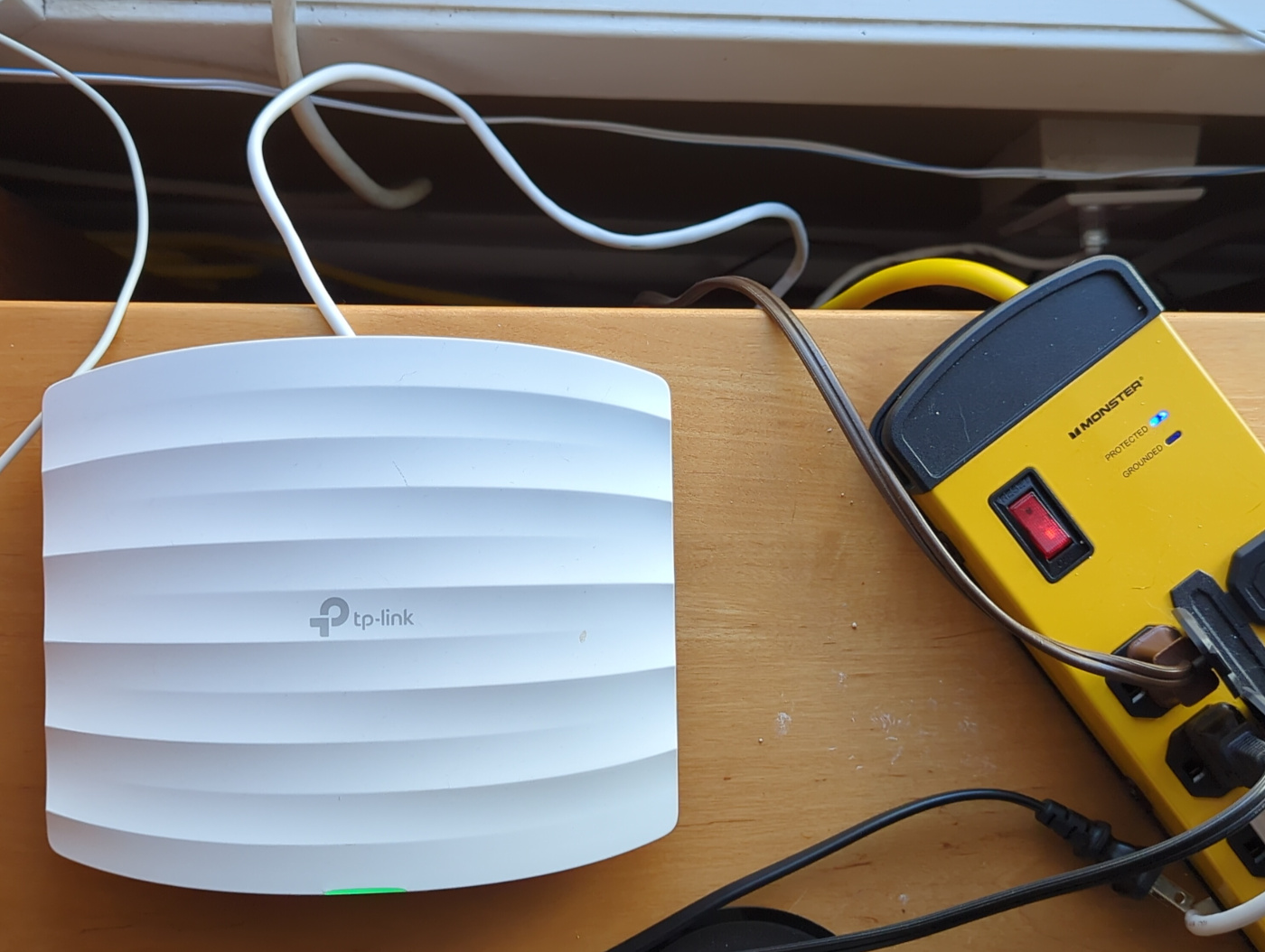 a white TP-Link device sitting on a wooden table next to a window, plugged into the wall by a white ethernet cable. it is not plugged into the power strip next to it.